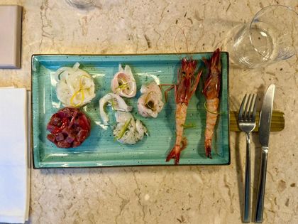 Piatto di spaghetti con pangrattato, pesce e basilico su un piatto blu. Tavola apparecchiata con posate e tovagliolo a quadretti.