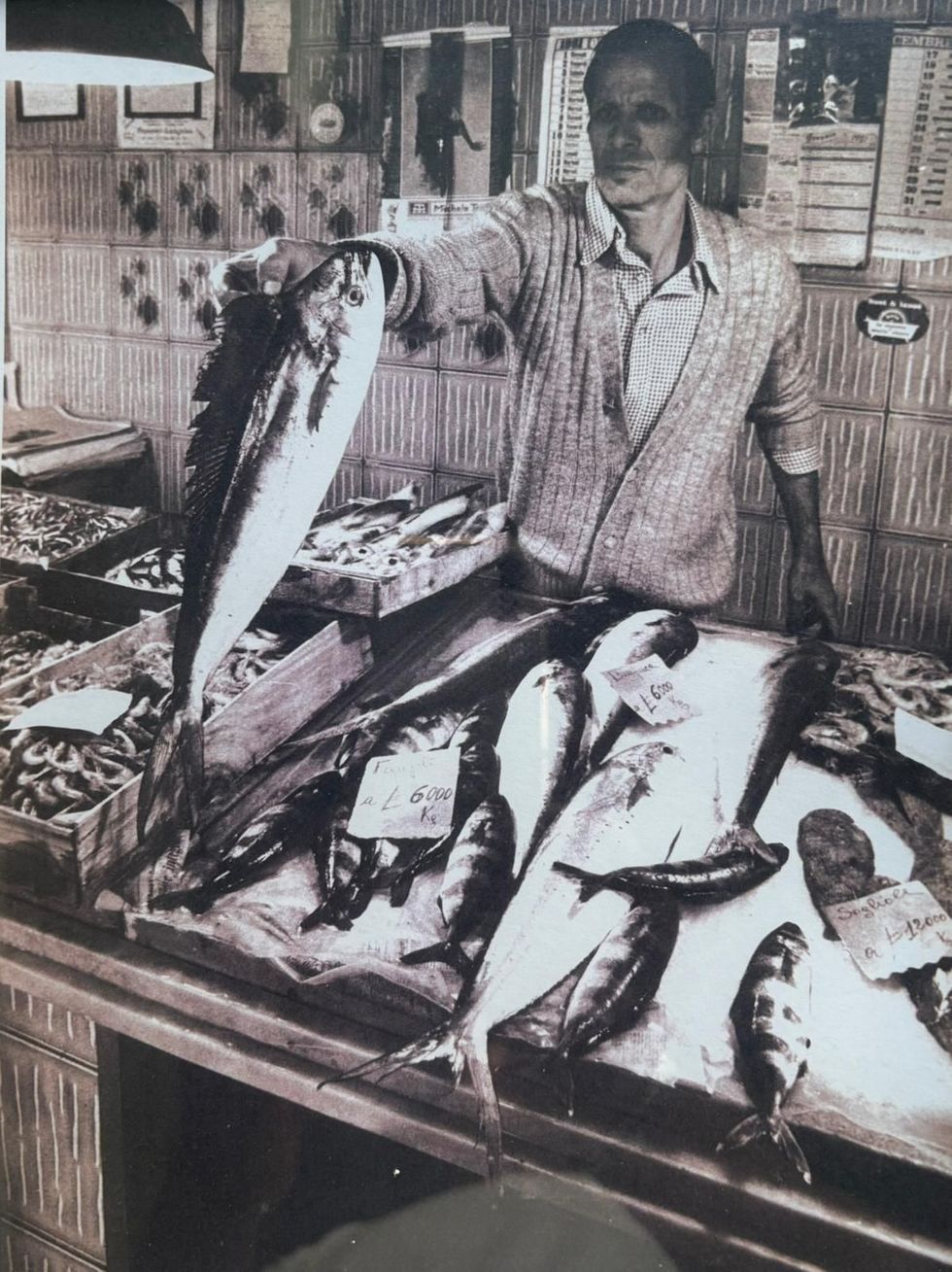 Un pescivendolo tiene in mano un grosso pesce, in piedi dietro un bancone che espone vari frutti di mare in un mercato.