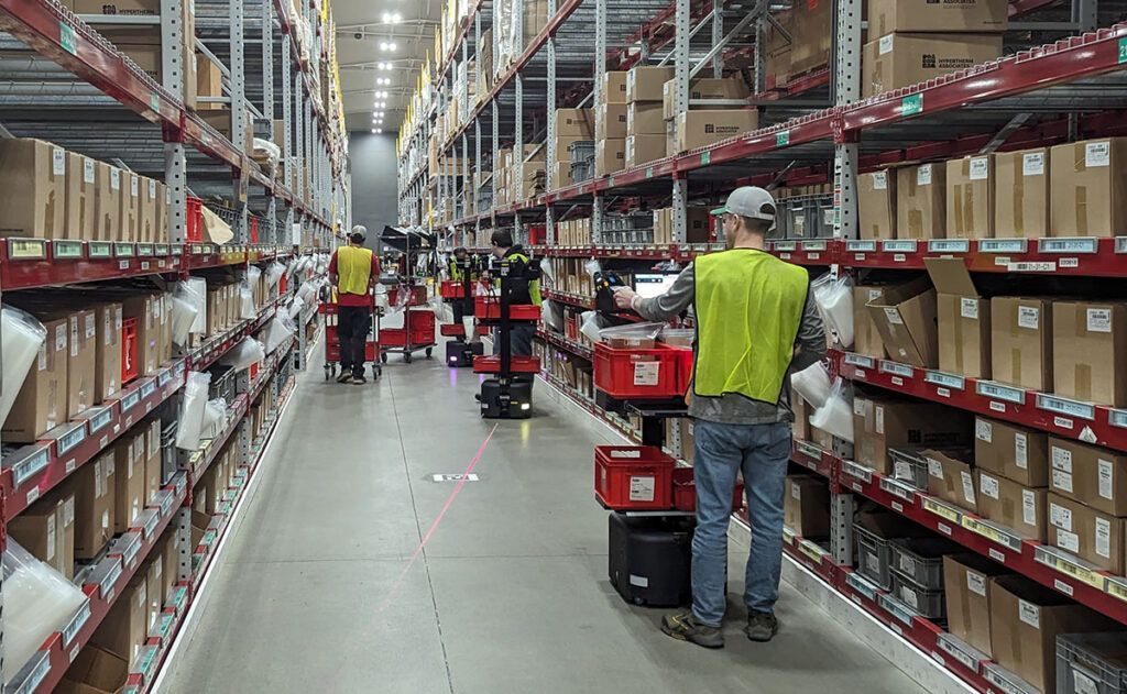 Warehouse aisle with workers stocking red carts beside tall shelves of boxed inventory