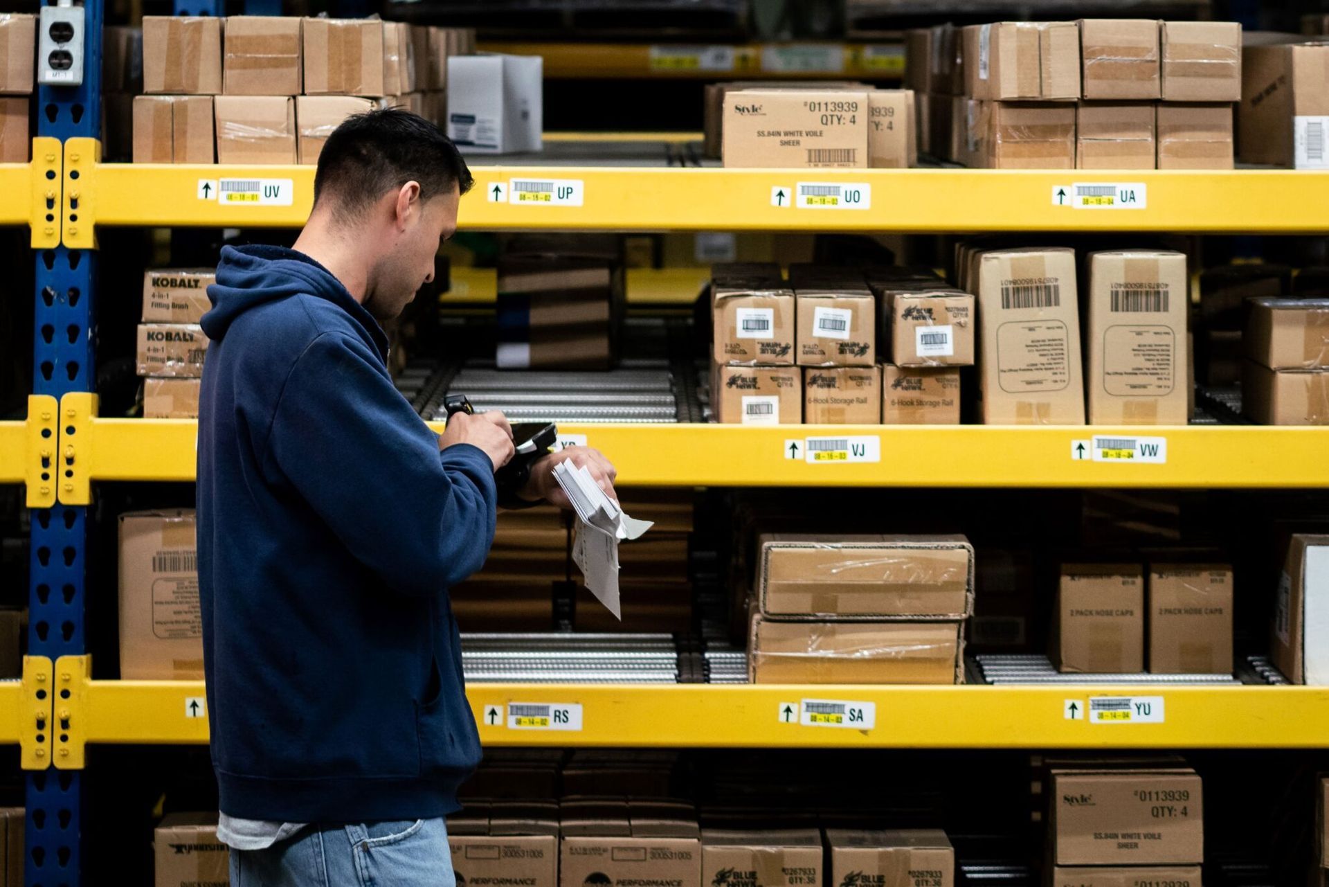 Worker in blue jacket inspecting packages on warehouse shelves with yellow racks