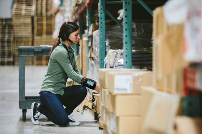 Warehouse worker kneeling by shelves, inspecting boxes in a storage aisle.