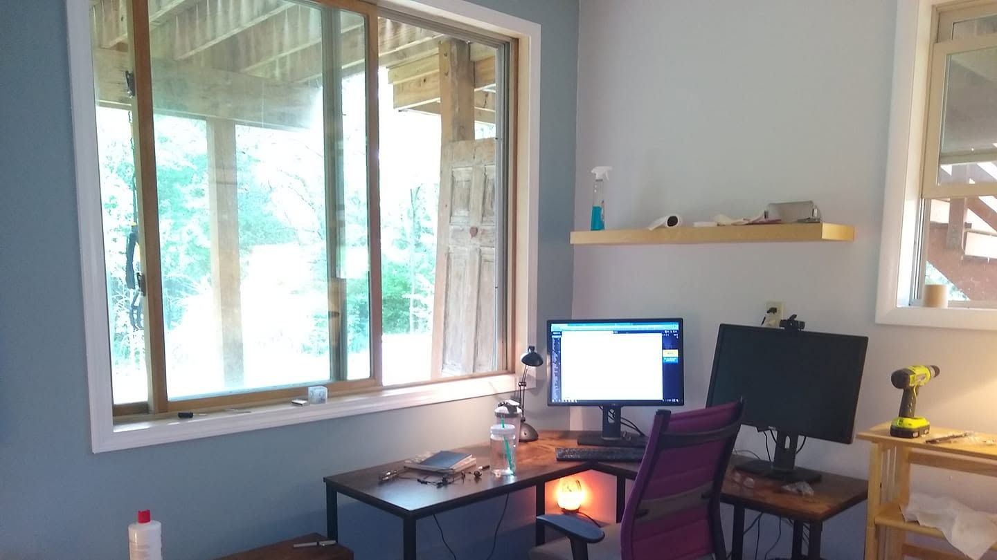 Home office with desk, computer monitor, purple chair, and large window overlooking greenery.