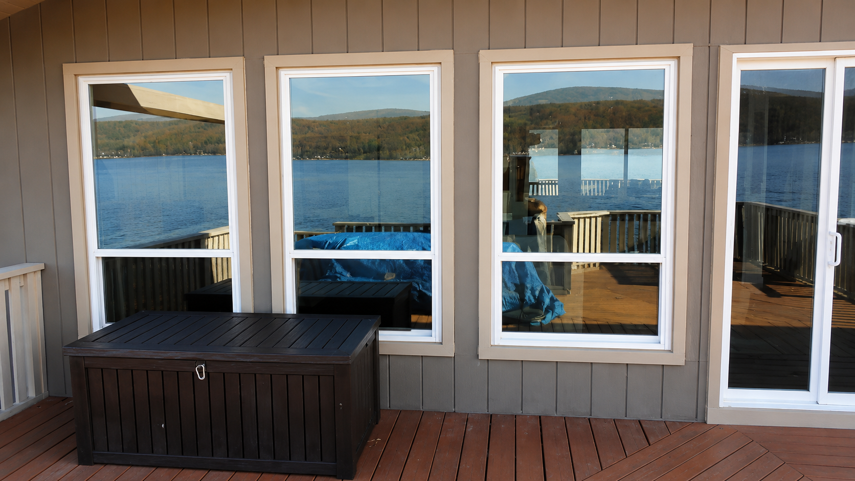 Wooden porch with large windows overlooking a lake and distant hills, with a dark storage bench in front.