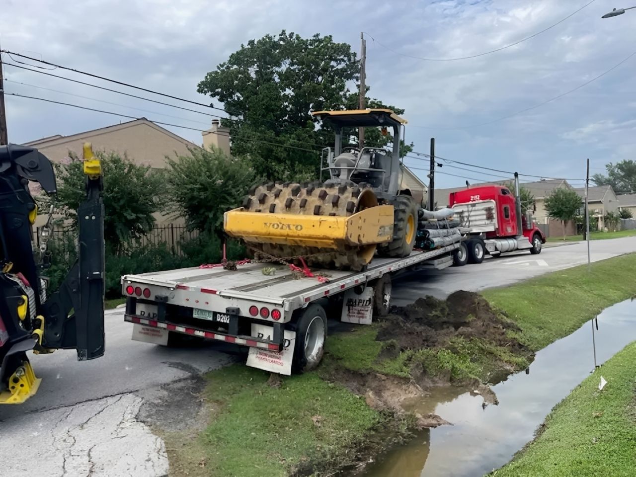 Bulldozer loaded on a flatbed truck beside a roadside ditch, with workers nearby on a cloudy day