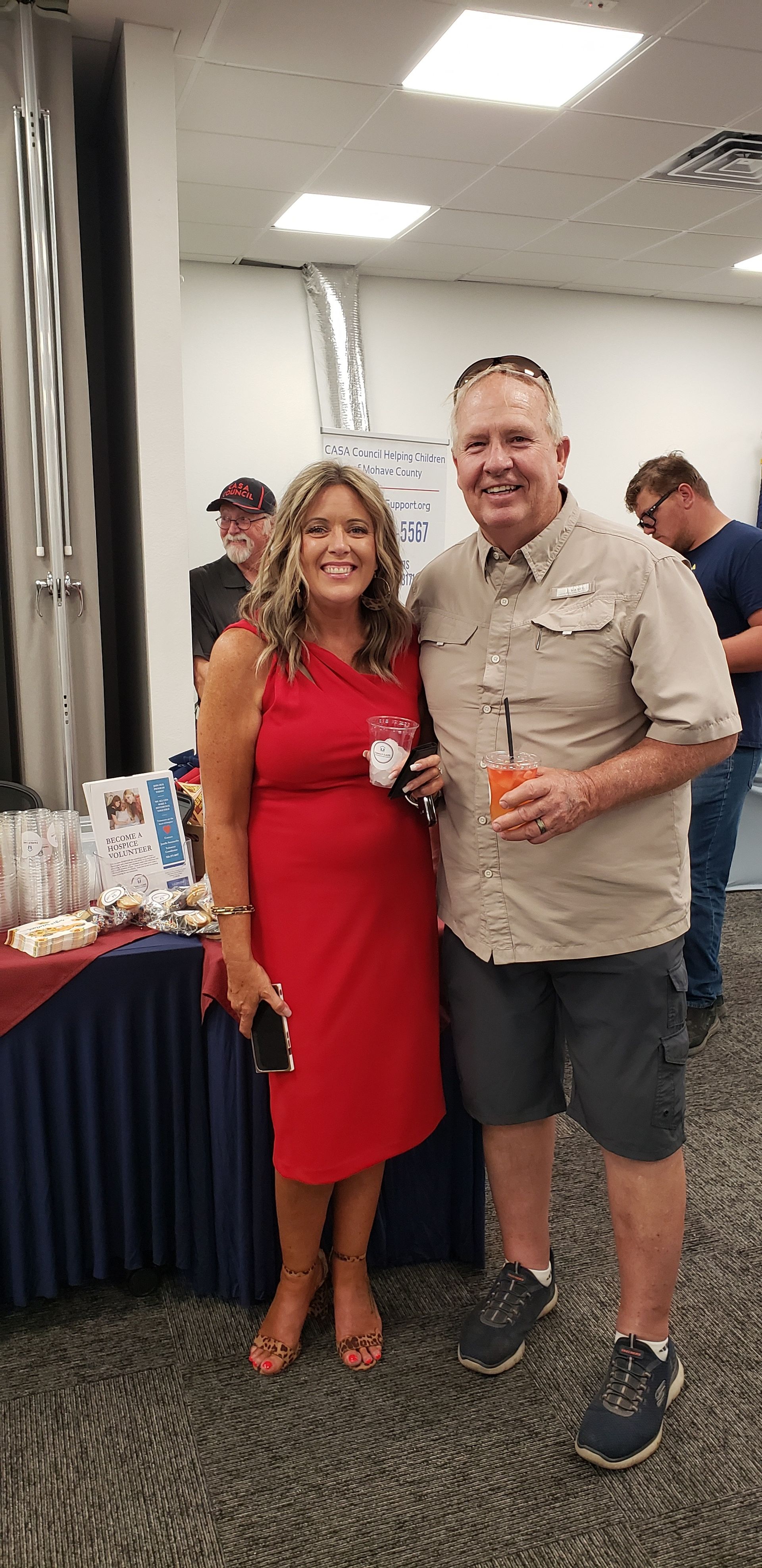Two people smiling, posing together. Woman in red dress, man in khaki shirt and shorts. Party setting.