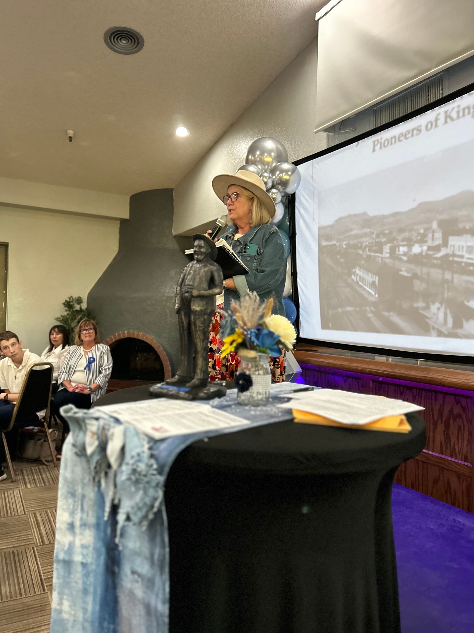 Woman speaking at podium with statue, projection screen behind her, in a room.