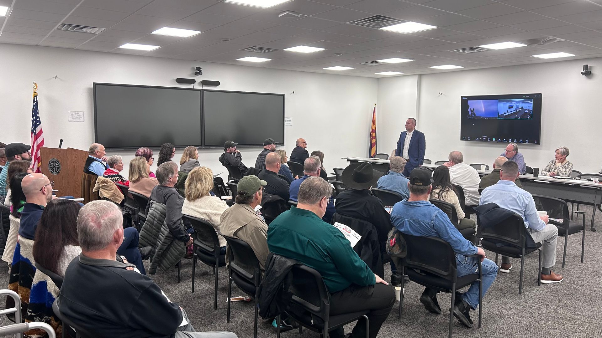 Meeting room with a speaker addressing an audience seated in chairs; multiple screens, and people at a table.