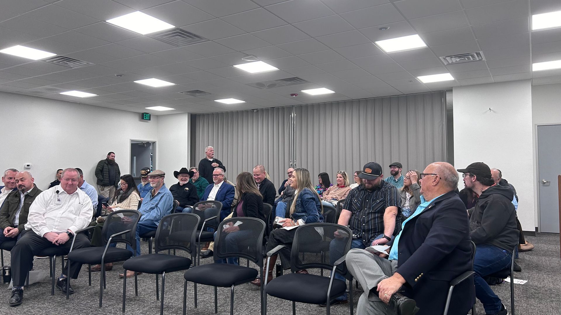 Audience in a meeting room, some seated, some standing, listening to a speaker. Neutral lighting.