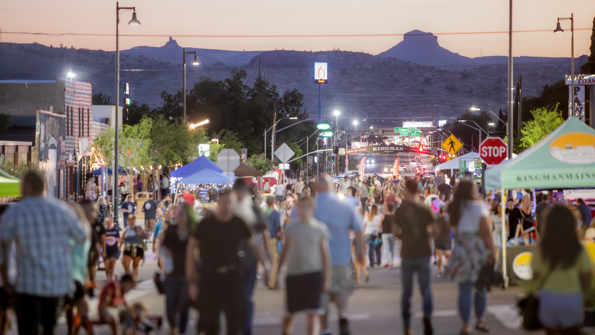Crowd of people on a street lined with shops and tents at dusk, with a mountain in the background.