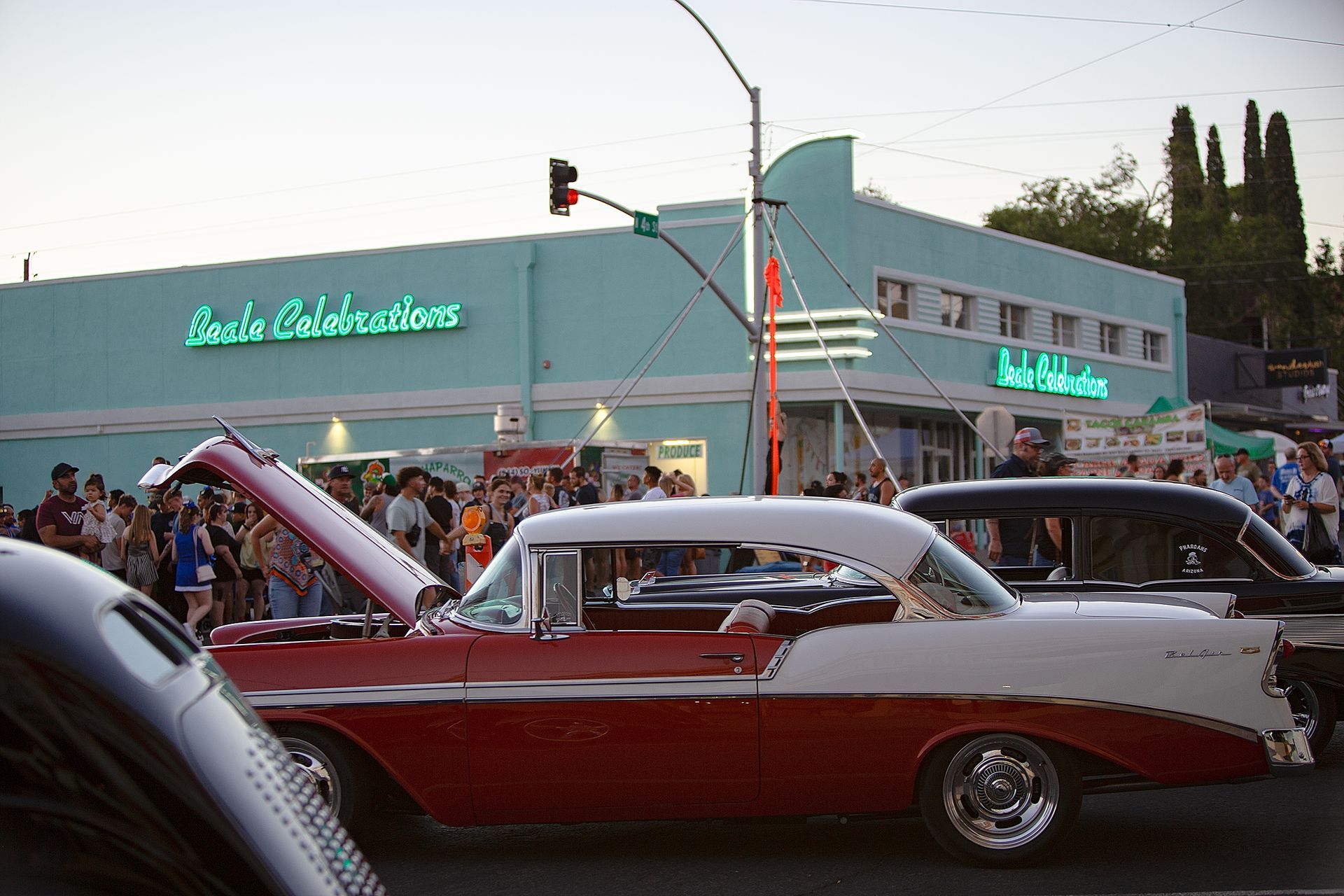 Classic cars parked on a street with a light blue art deco building in the background. A crowd of people stand on the sidewalk.