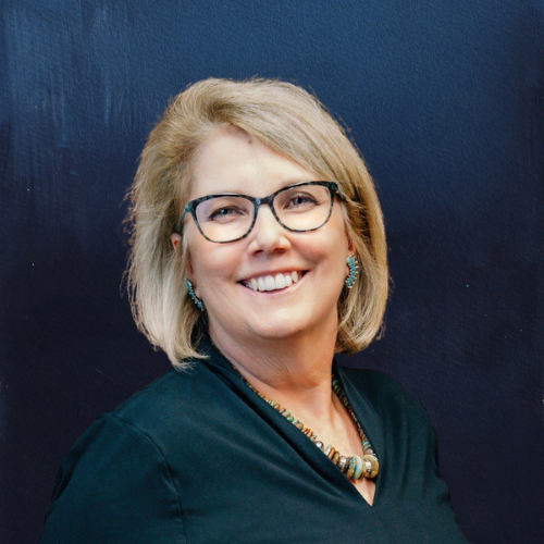 Woman with glasses smiles; wearing teal earrings, necklace, and a dark green shirt; dark blue background.