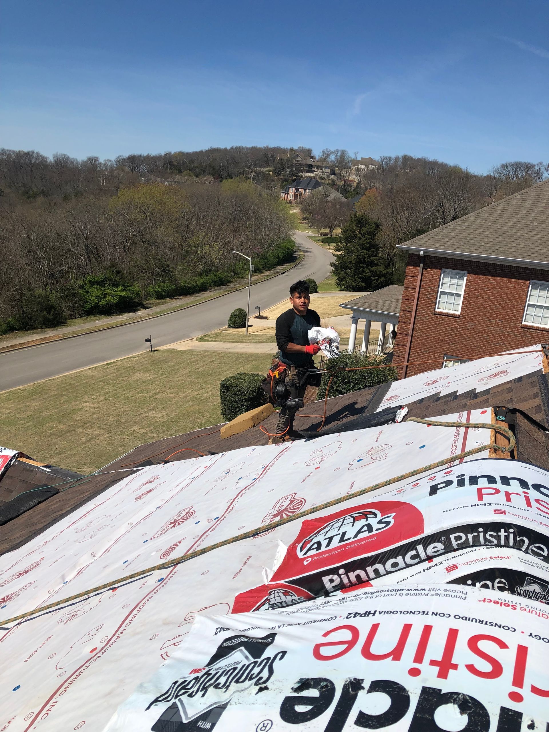 A roofer installs dark shingles on a residential roof, seen from a low-angle close-up.