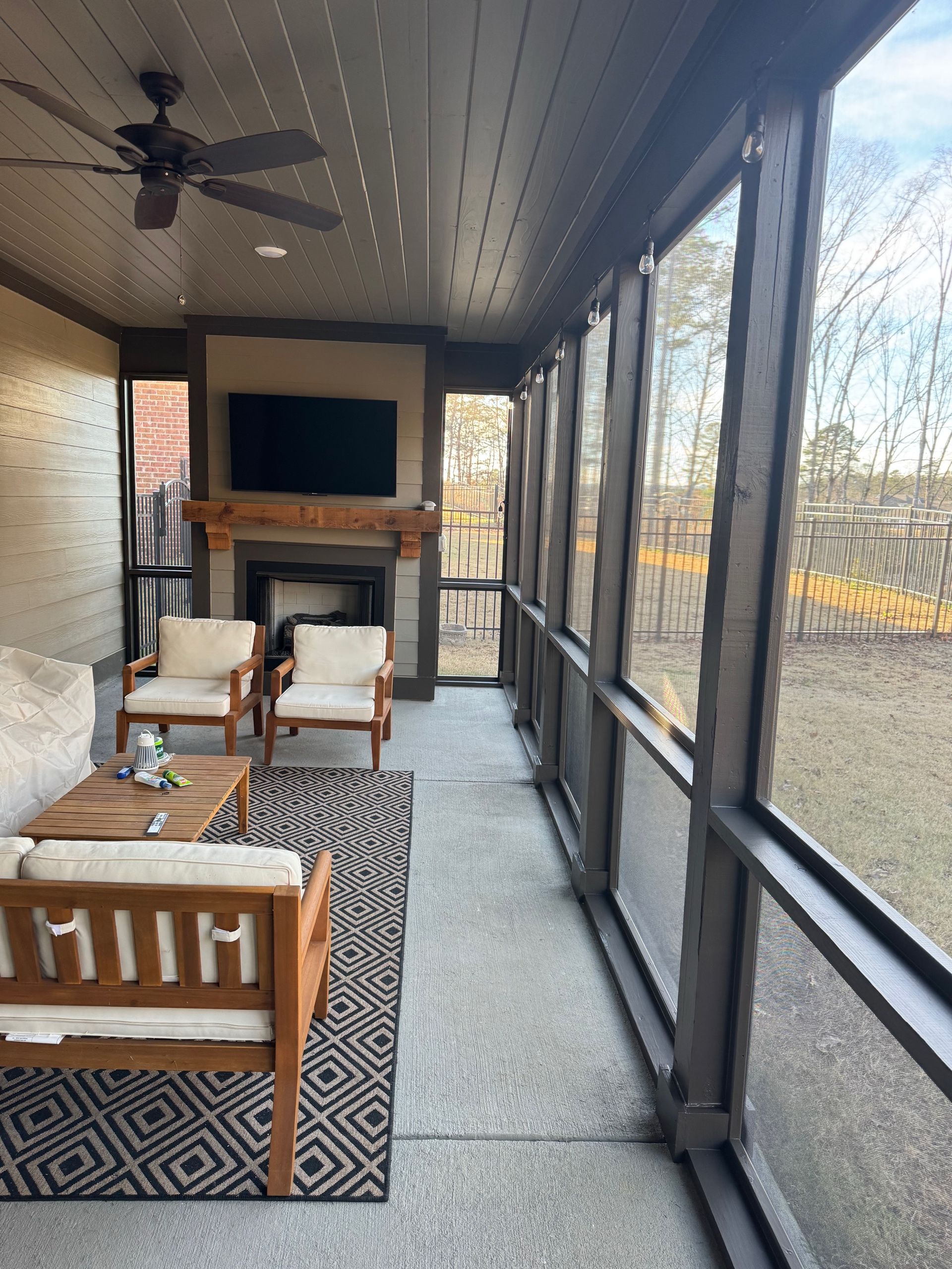 A furnished covered patio with a fireplace, wall-mounted television, and floor-to-ceiling screens overlooking a yard.