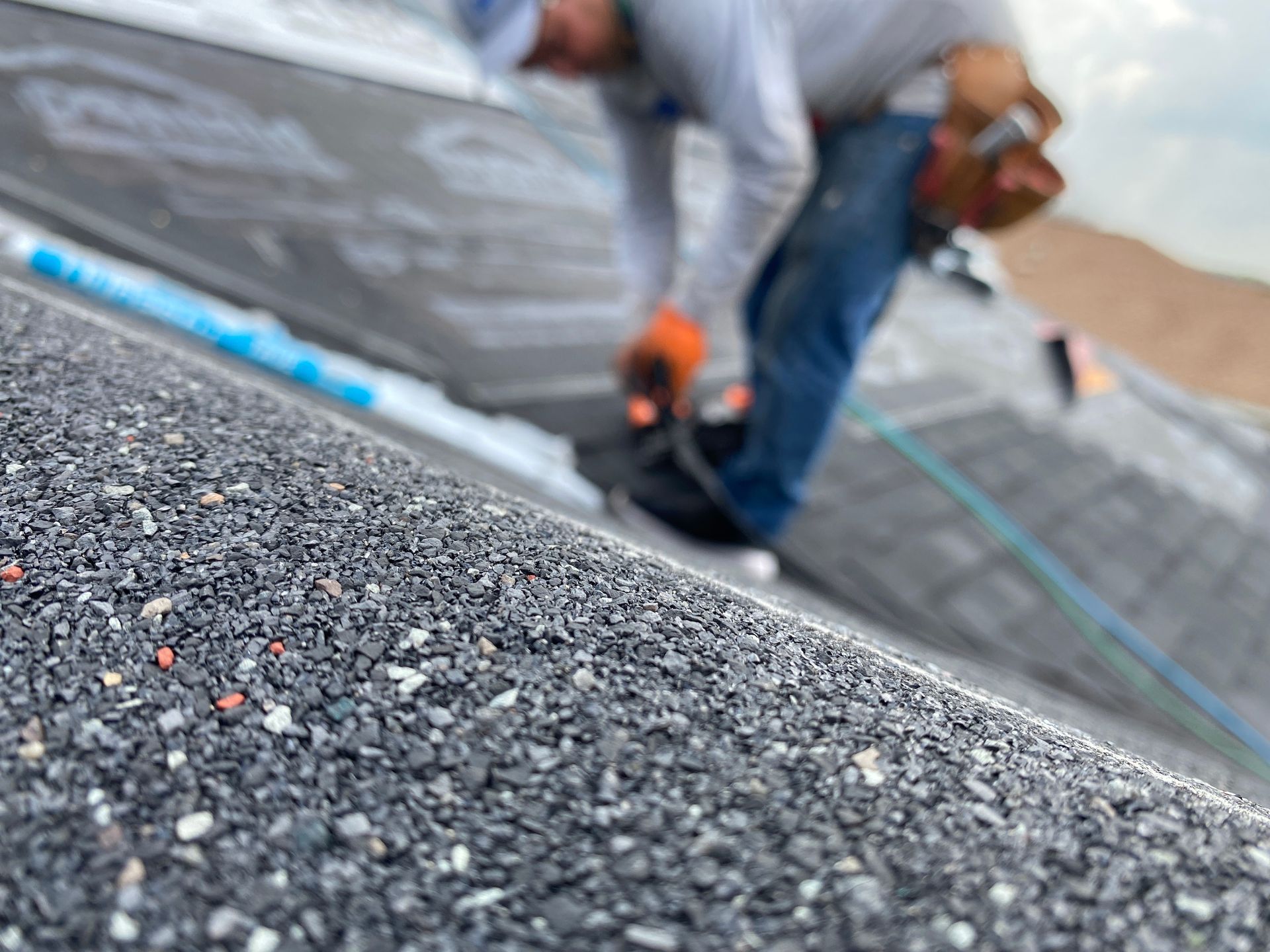 A roofer installs dark shingles on a residential roof, seen from a low-angle close-up.