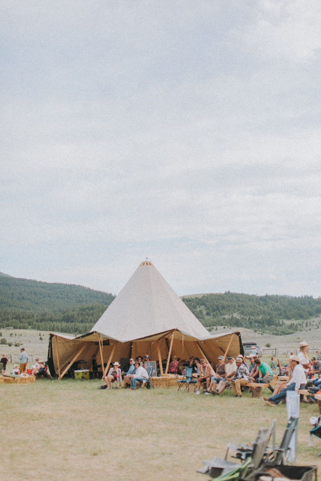 Outdoor festival campsite with a large white teepee tent and people gathered around under a cloudy sky