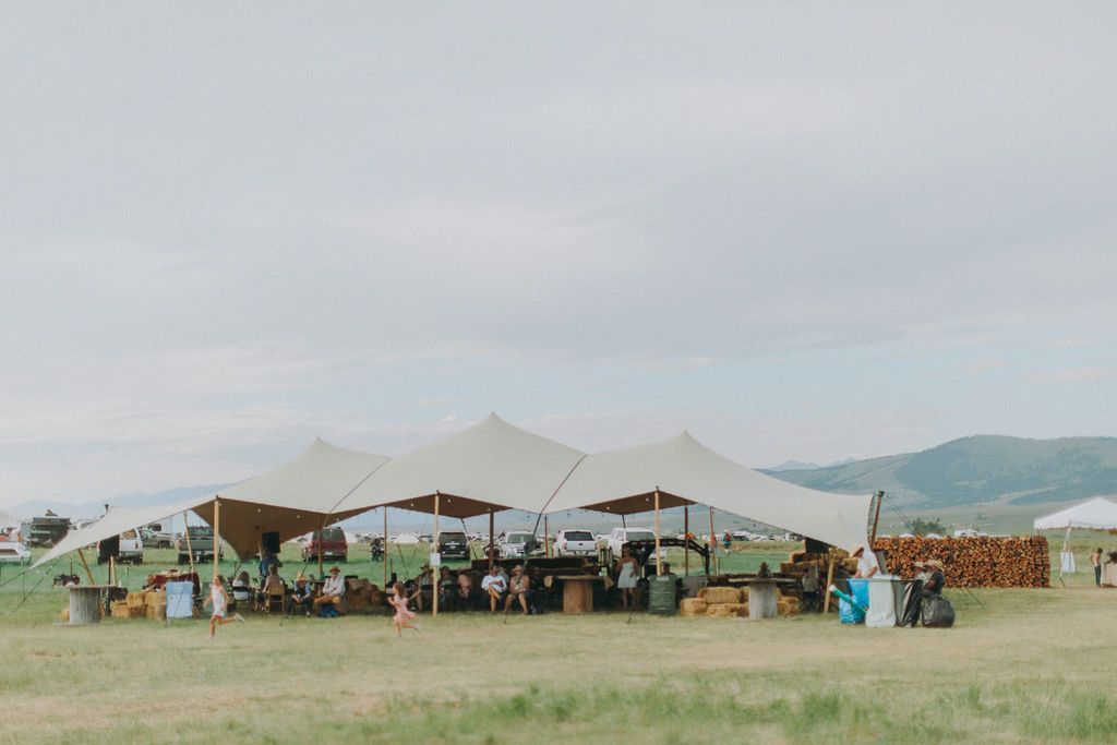 Open-air market tents on a grassy field with people and stacked wood under an overcast sky