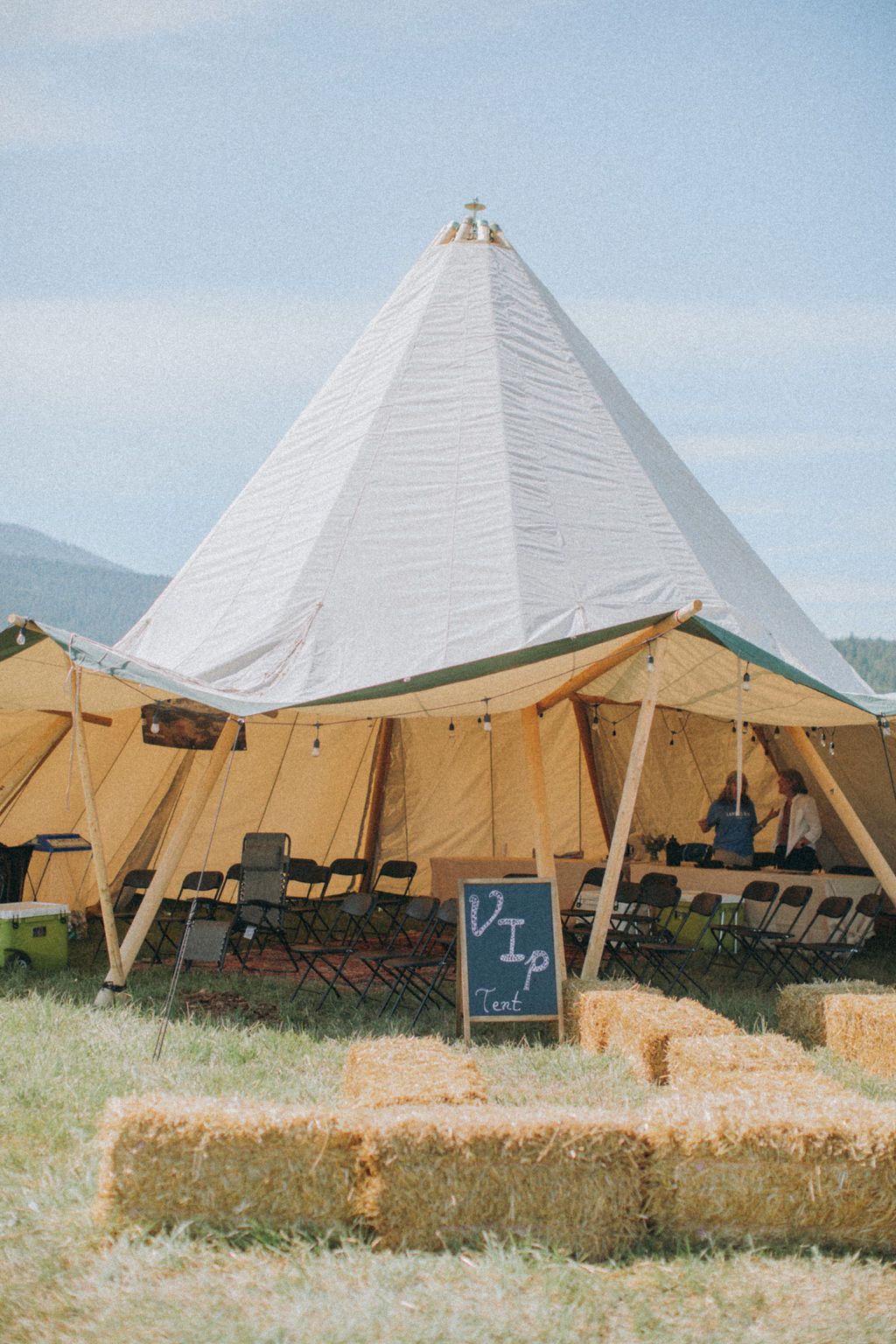 White tent pavilion with hay bale seating and a small chalkboard sign in a grassy outdoor field