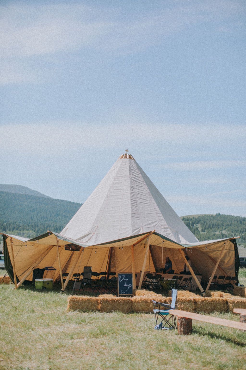 White tent-like pavilion with tan canopy in a grassy outdoor field, with hills and blue sky in the background