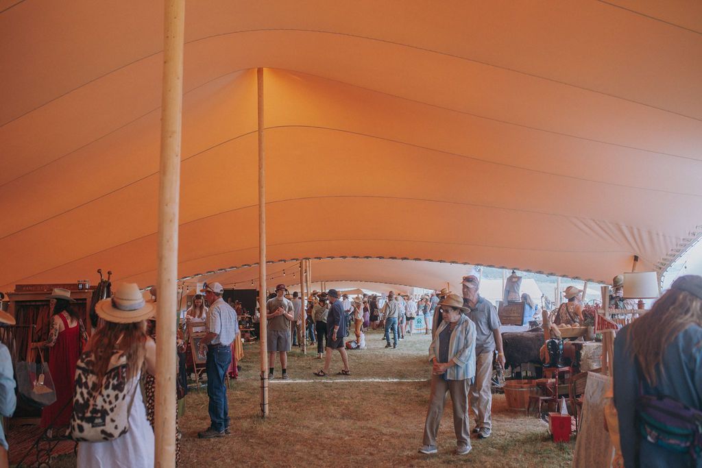 Inside a large orange tent, people browse and socialize at an indoor market or fair.