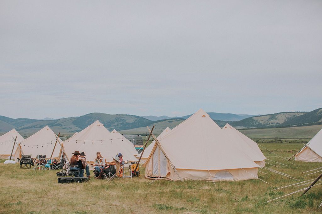 Several beige tents in a grassy field with people gathered outside under a cloudy sky.