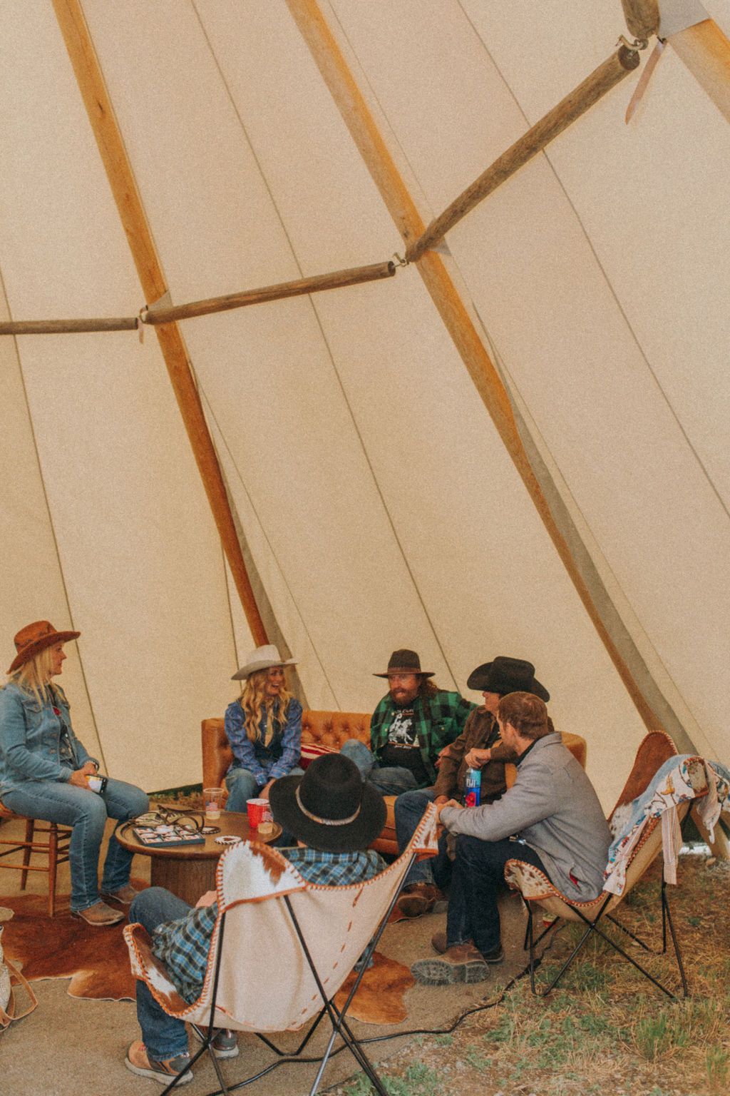 People seated in folding chairs around a campfire inside a large canvas tent.