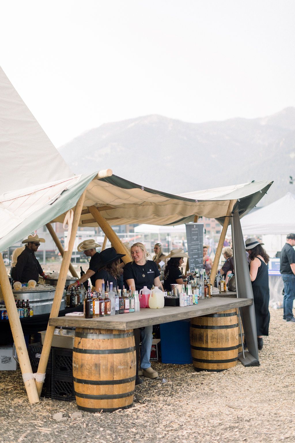 Outdoor wooden drink stall under a canopy, with people chatting at a festival in a mountain setting
