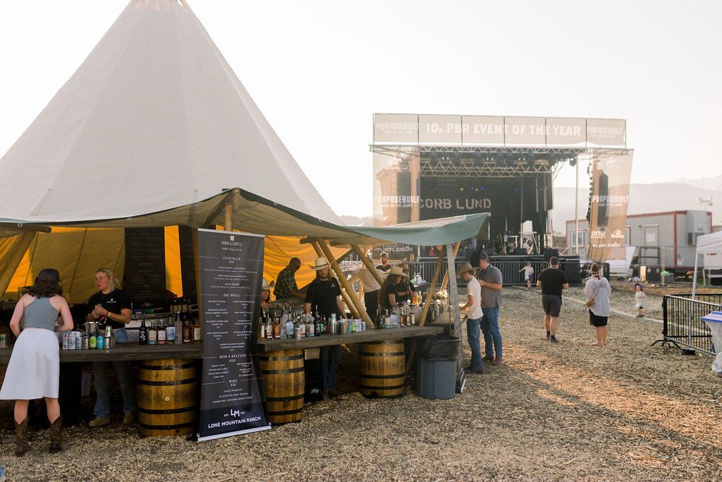 Outdoor festival tent with people at a bar, and a stage in the background under a hazy sky