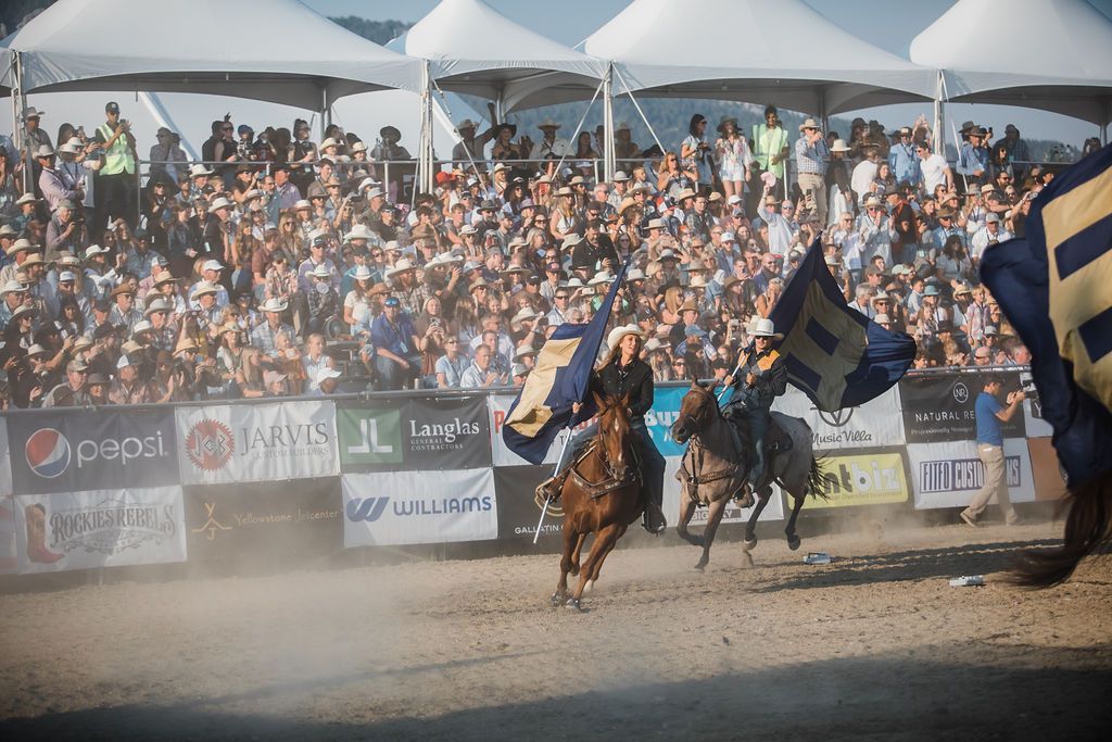 Horse riders with flags perform before a cheering crowd at a dusty outdoor arena.