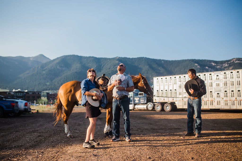 People standing with horses in a dusty field, with mountains and a trailer in the background.