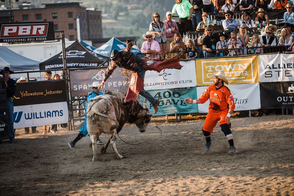 Bull rider thrown from a bucking bull in a rodeo arena, with a clown nearby and spectators watching.