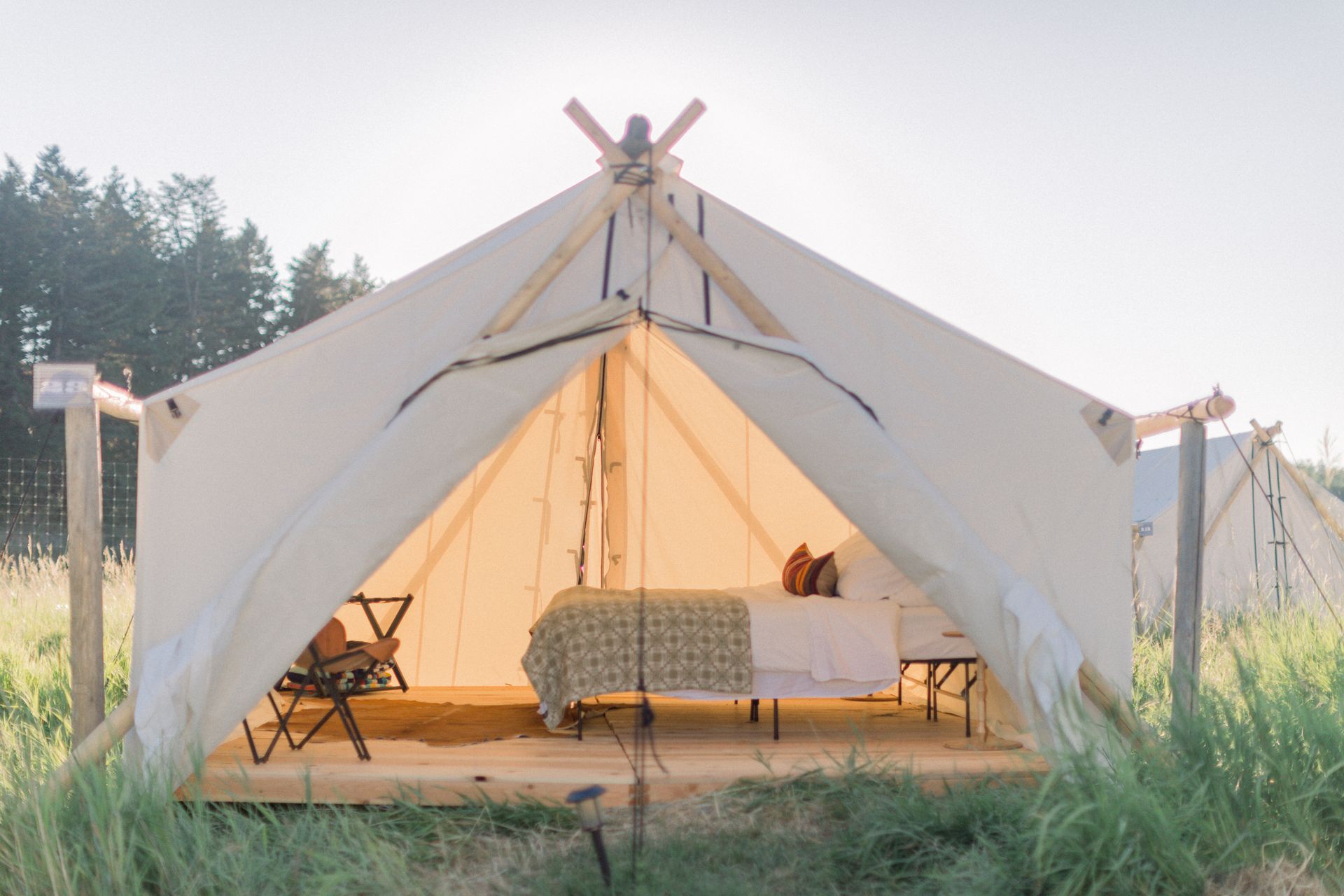 Open white canvas tent with bed and chairs in a grassy outdoor field at sunset