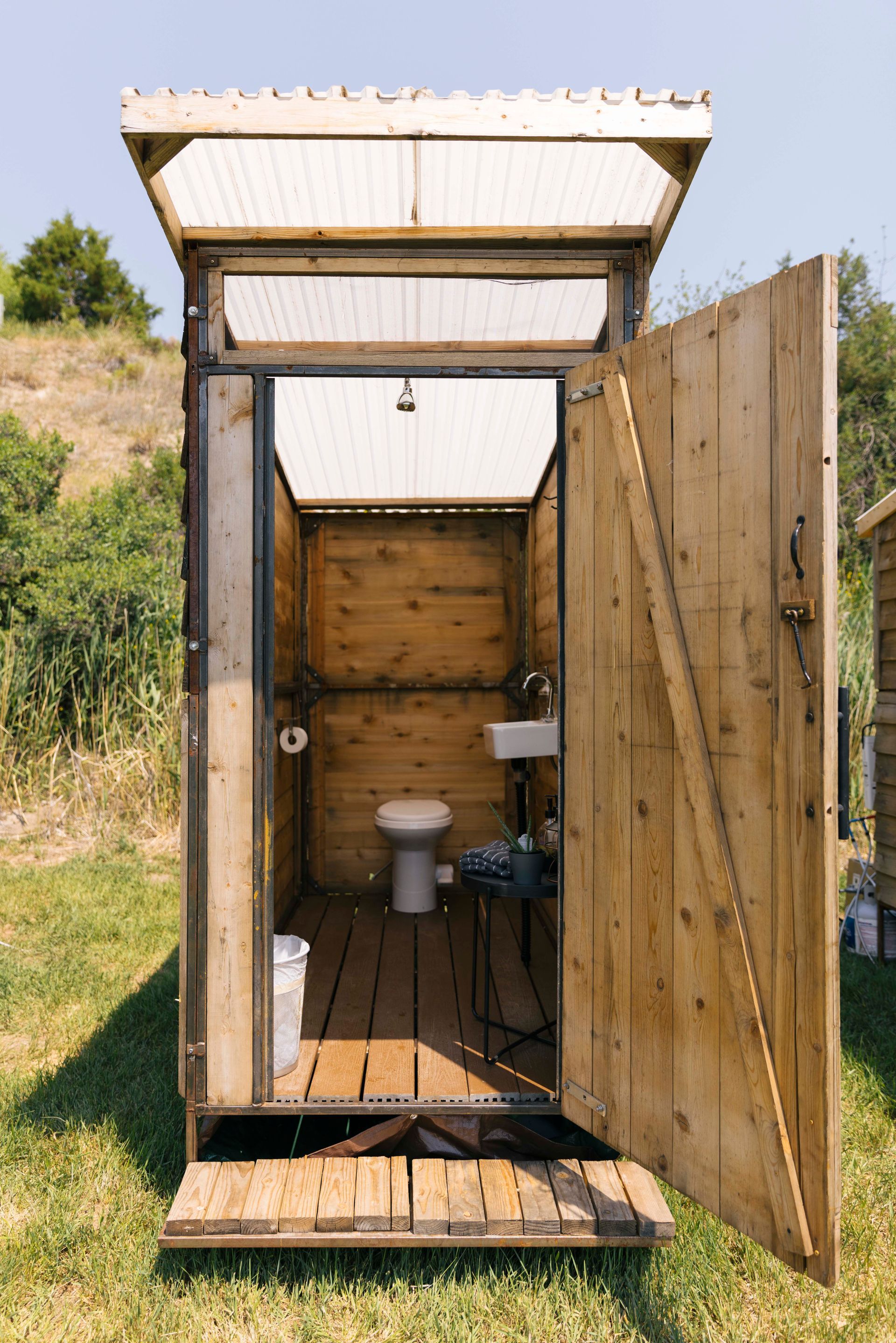 Outdoor wooden toilet stall with open door, visible toilet and sink inside