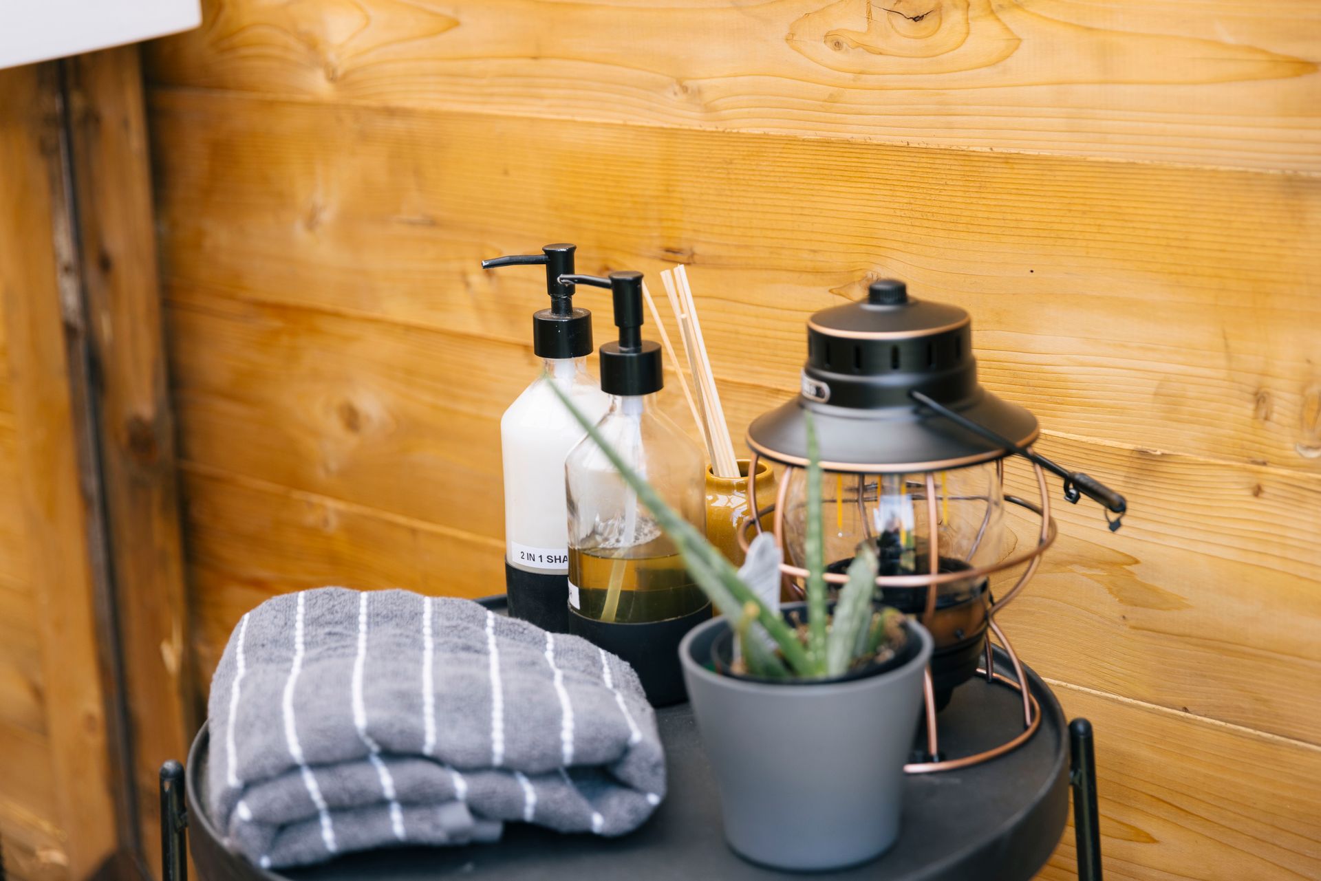 Striped towel, potted plant, spray bottles, and lantern on a black table against a wooden wall
