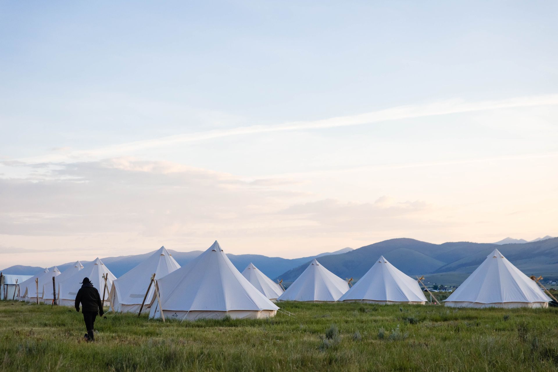 White tents in a grassy field at dusk, with a person walking in the foreground and blue hills in the distance