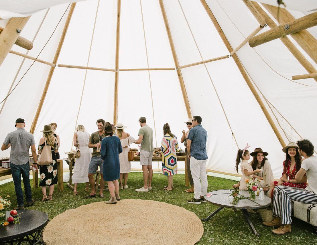 People socializing inside a white teepee tent with grass flooring and woven seating areas