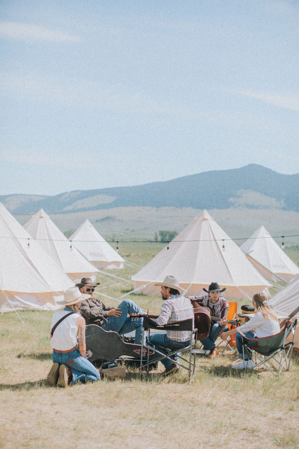 People camping and sitting around a table near white tents in a grassy field with mountains in the background