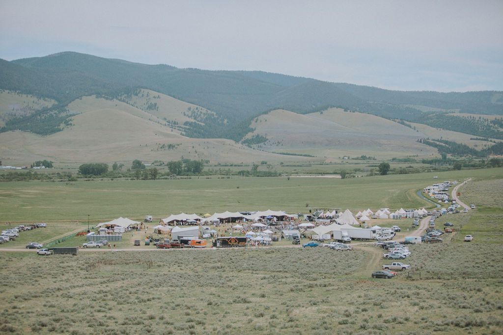 Large outdoor encampment in a grassy valley with tents and vehicles, backed by rolling hills under a hazy sky
