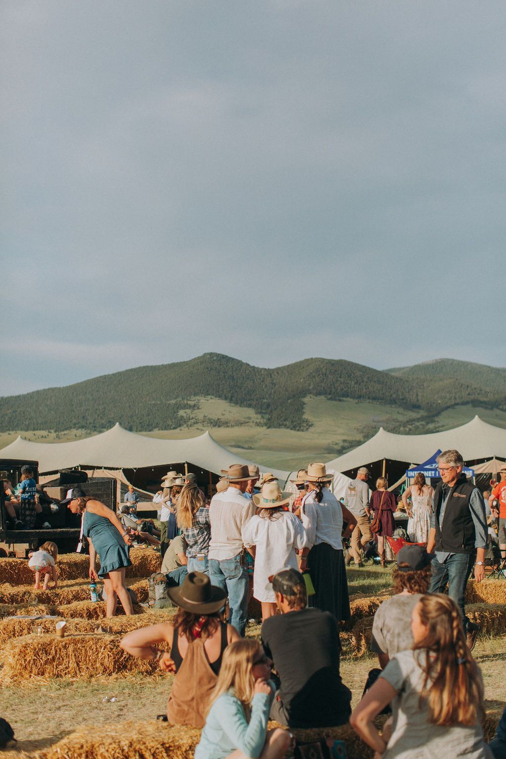 Outdoor festival crowd among straw bales and tents, with hills under a cloudy sky in the background