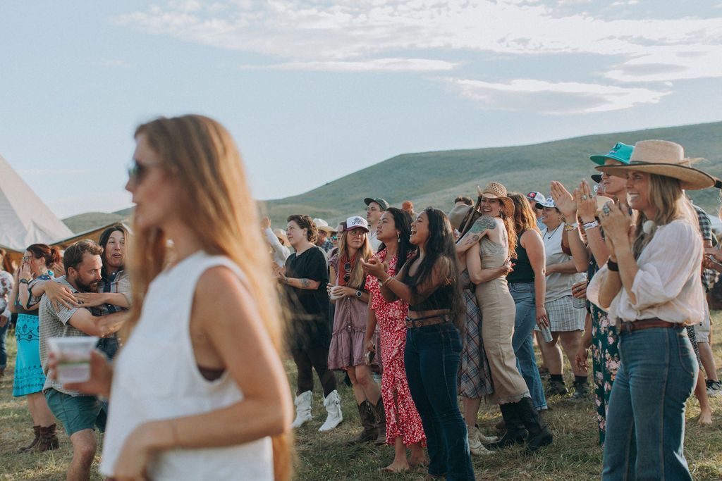 Crowd dancing outdoors at a music festival, with a woman in a white shirt in the foreground and hills behind.