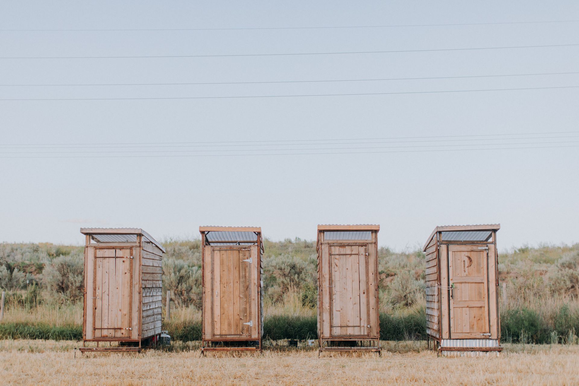 Four wooden outhouses lined up in a grassy field under a pale sky.