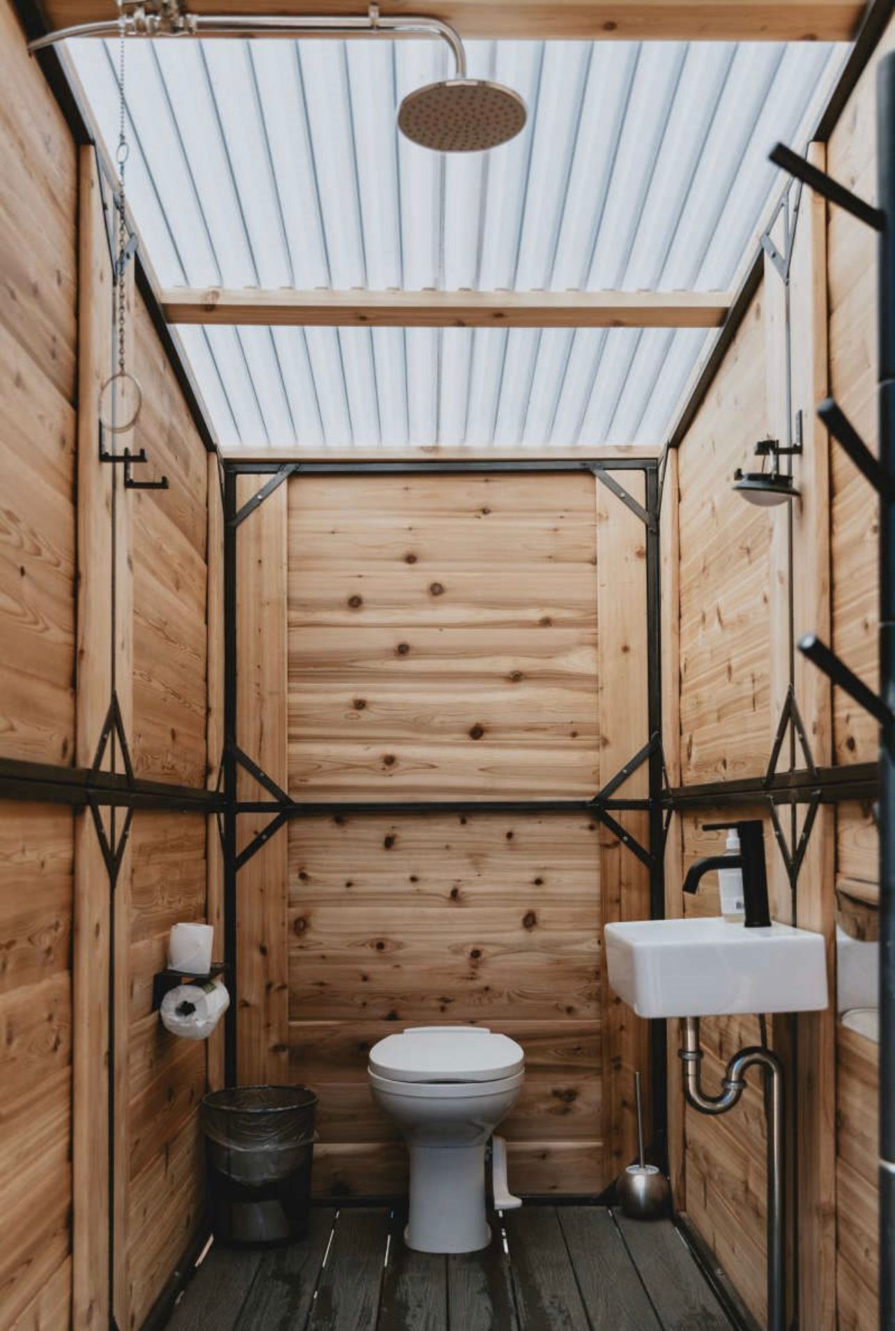 Rustic outdoor bathroom with wooden walls, toilet, sink, and shower under a corrugated roof