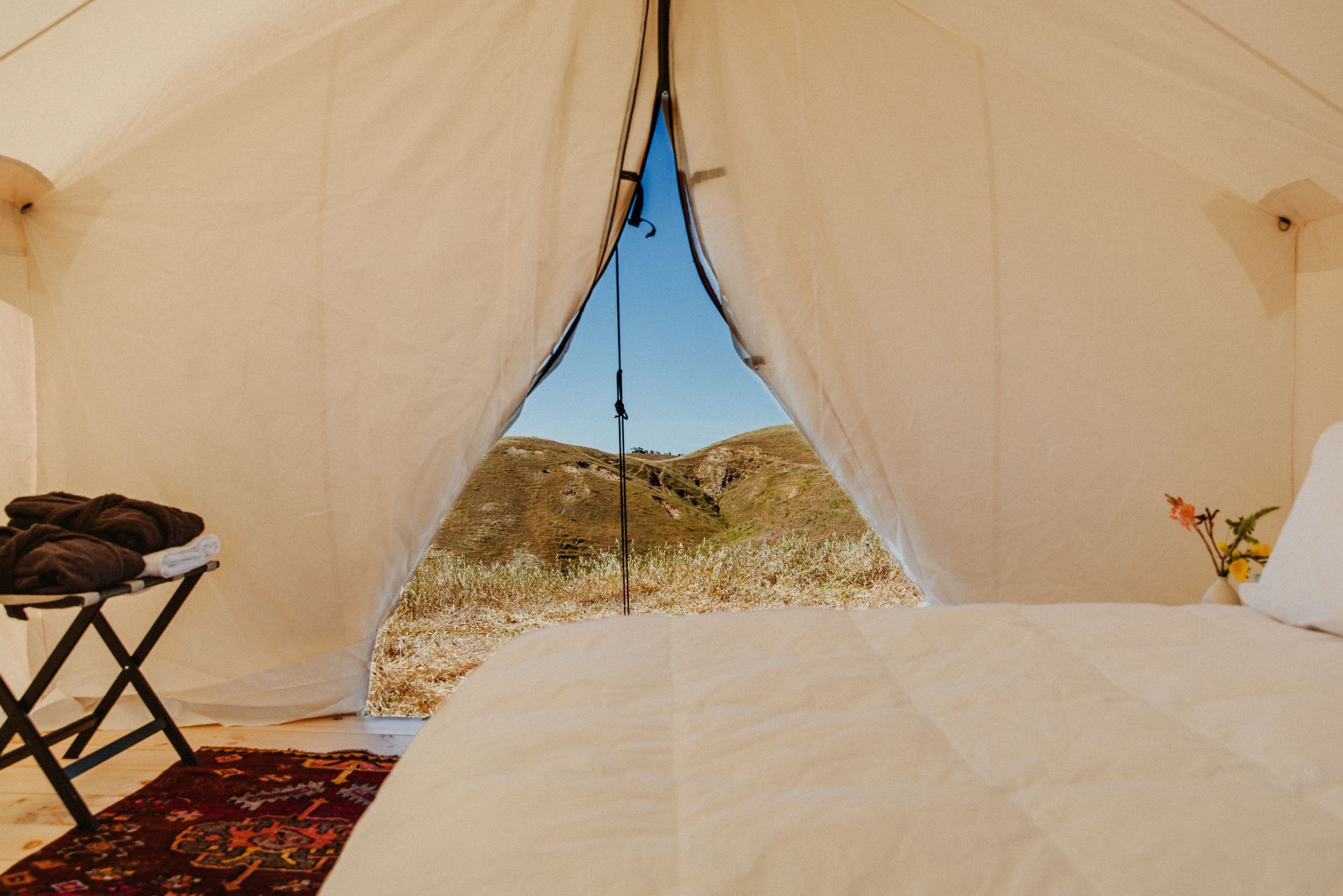 Inside a cream tent, a bed faces an open doorway with a view of hills and blue sky.