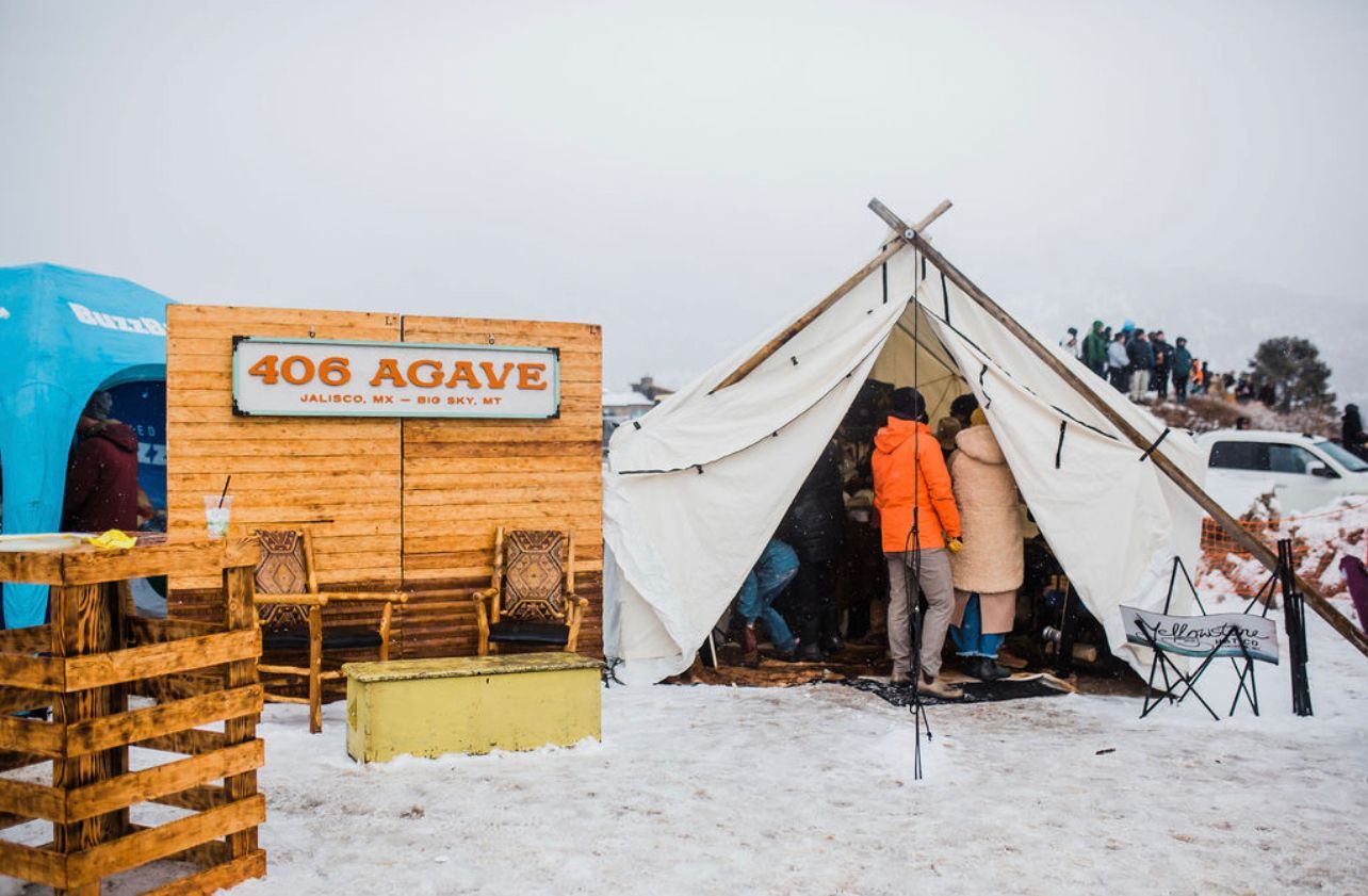 Snowy campsite with a white tent and orange-clad person beside a “406 AGAVE” wooden stall