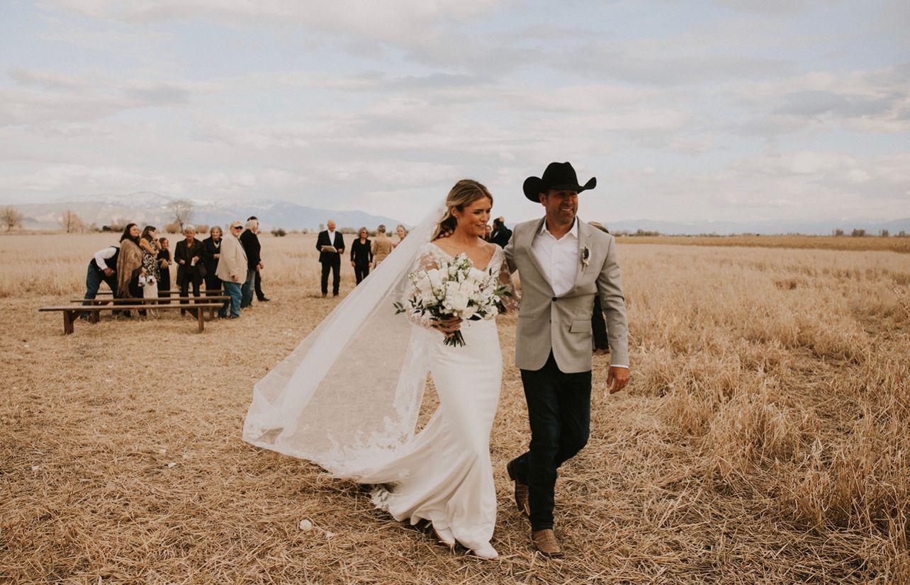 Bride and groom walking through a wheat field with wedding guests in the background