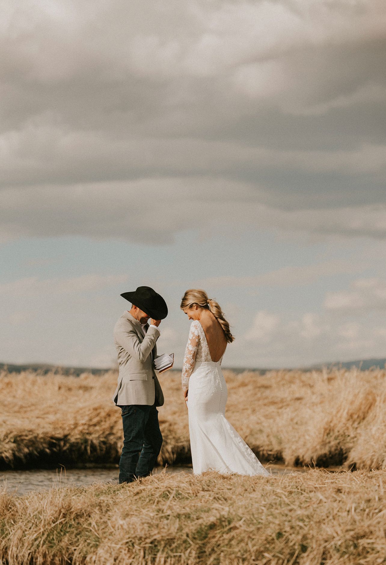 Couple standing in a grassy field under cloudy sky, dressed in wedding attire, holding hands