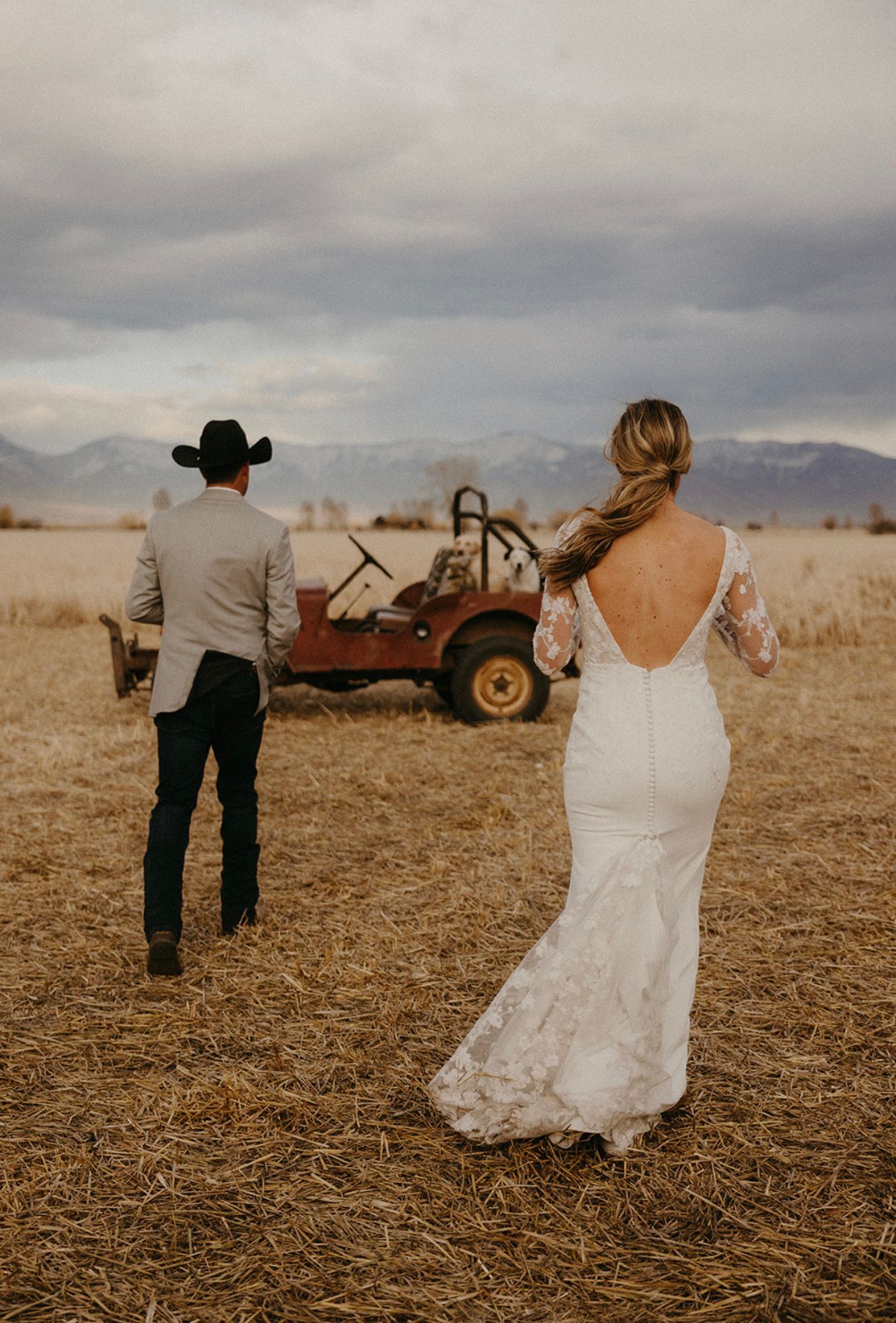 Bride and cowboy walking toward a red off-road buggy in a dry field under cloudy skies
