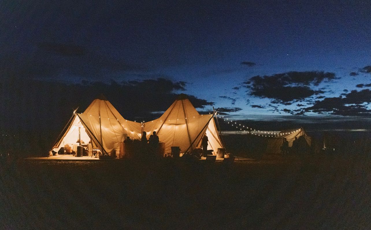 Lit tents glowing in a dark field at dusk under a blue sky