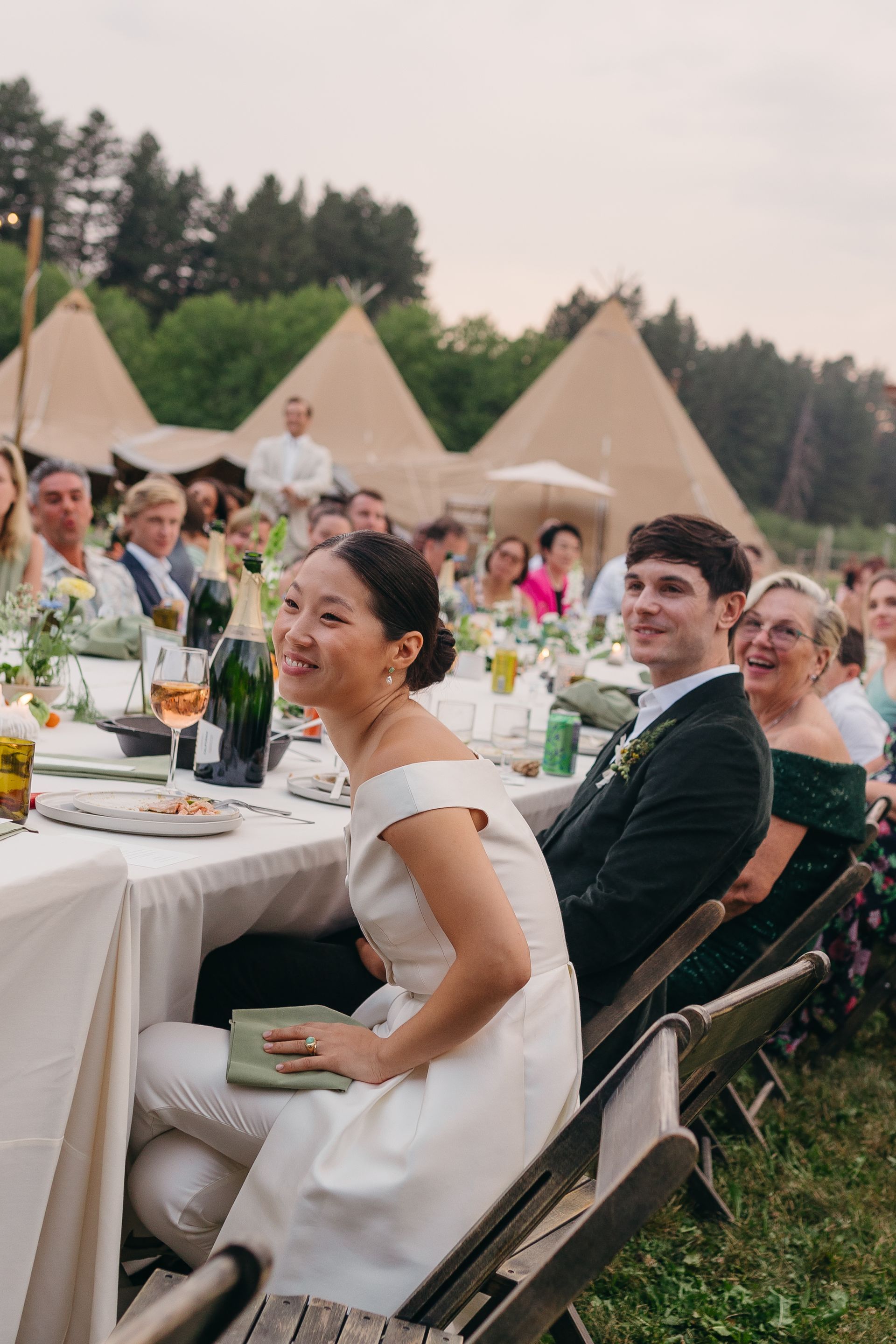 Outdoor wedding reception with guests seated at long tables under tents, a bride smiling in the foreground.