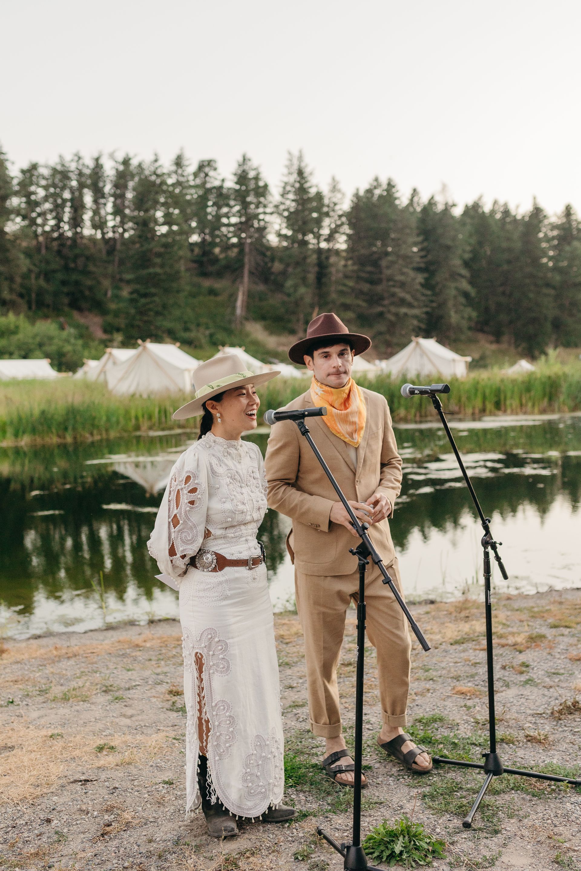 Two people in vintage outfits stand by microphones beside a lake with tents and pine trees behind them.