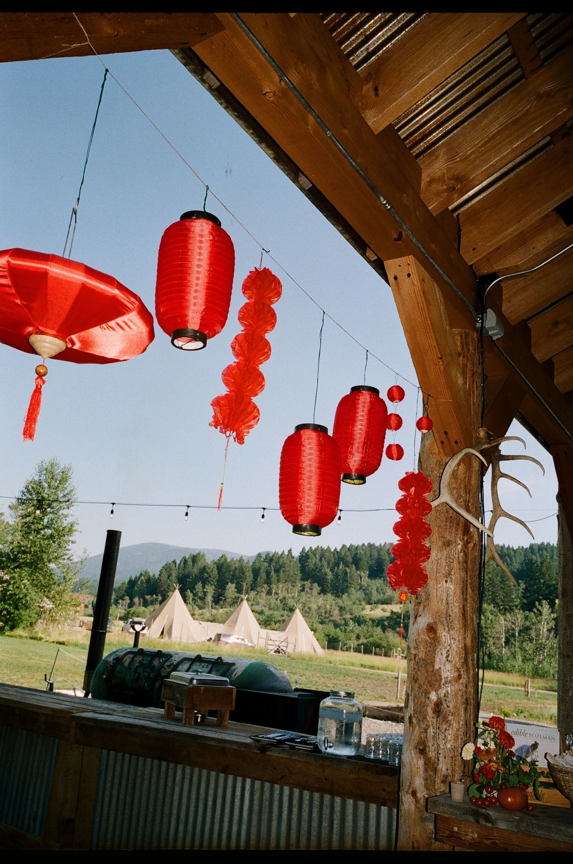 Red lanterns hang from a wooden porch overlooking a sunny outdoor field with tents.